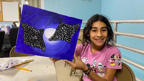 A student holding a canvas with a painting of spotted eagle rays on a blue background.